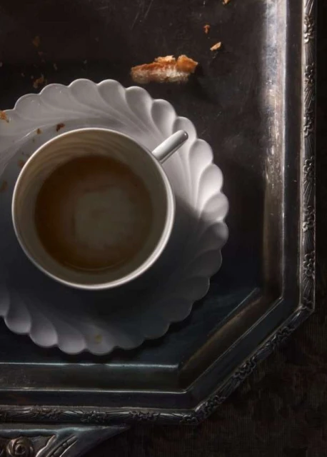 Overhead shot of a cup of coffee on a decorative plate and silver tray with pastry crumbs, creating a simple and elegant still life.