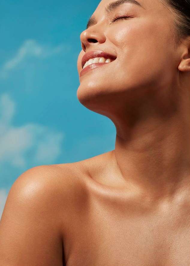 Close-up portrait of a smiling woman with glowing skin against a blue sky with clouds. She has her eyes closed and is facing upwards.