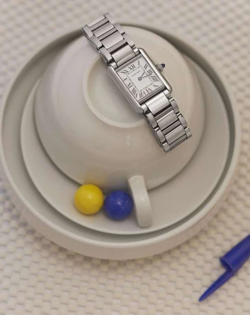 Elevated view of a silver Cartier Tank watch on a stack of white dishes with colorful decorative balls, creating a minimalist aesthetic.
