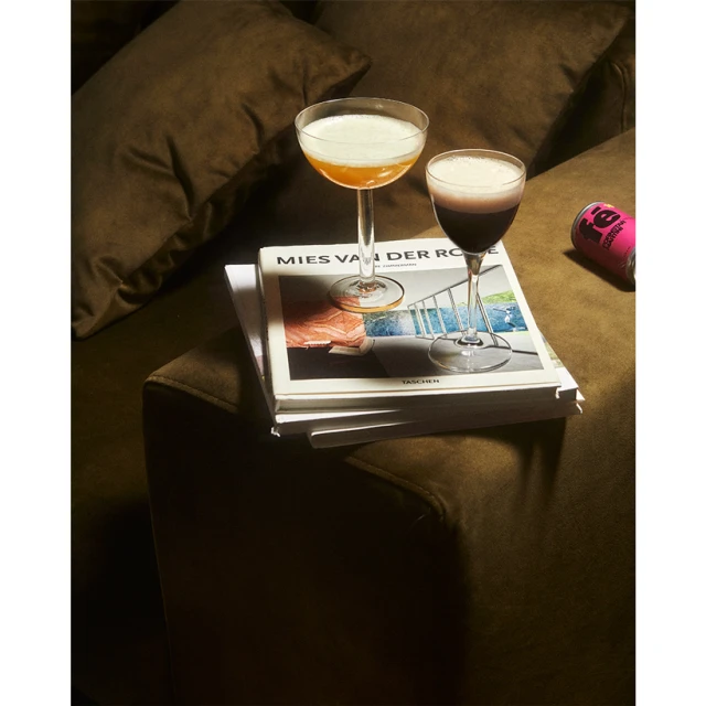 Two cocktail glasses on a stack of books atop a brown couch, with a pink can visible in the background, under studio lighting.