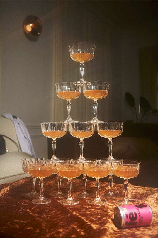 Tower of champagne coupe glasses filled with a golden beverage sits on a brown velvet table with a can of tea in the foreground.
