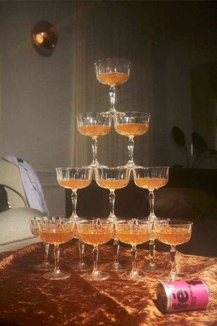 Tower of champagne coupe glasses filled with a golden beverage sits on a brown velvet table with a can of tea in the foreground.