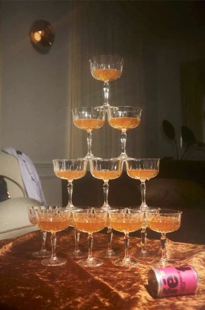 Tower of champagne coupe glasses filled with a golden beverage sits on a brown velvet table with a can of tea in the foreground.