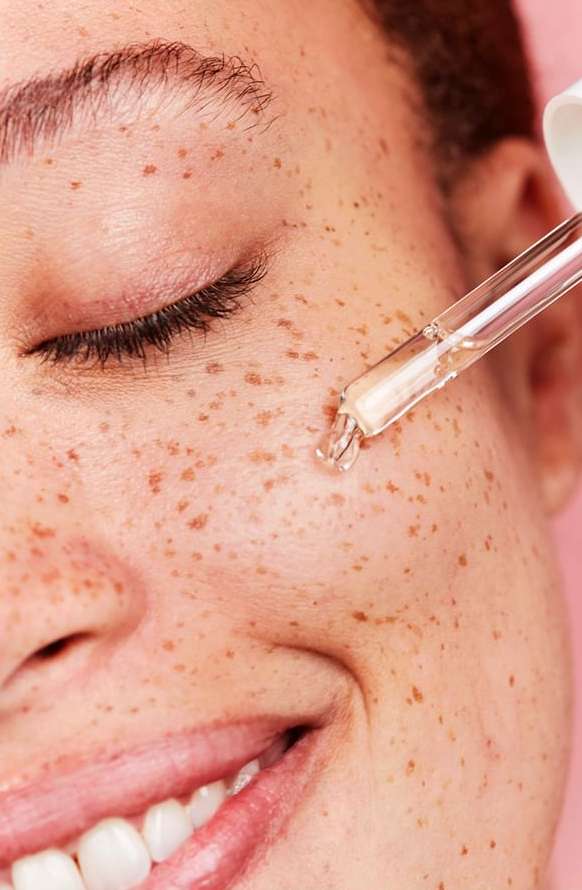 Close-up of a woman's face with freckles and a dropper applying liquid serum to her cheek in a beauty and skincare context.