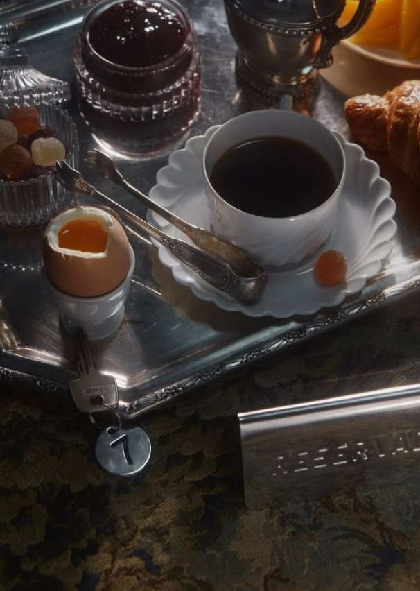 Breakfast tray with coffee, soft-boiled egg, jam, and pastries on a patterned surface, illuminated by natural daylight, creating a cozy mood.