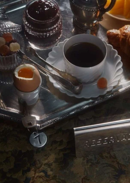 Breakfast tray with coffee, soft-boiled egg, jam, and pastries on a patterned surface, illuminated by natural daylight, creating a cozy mood.