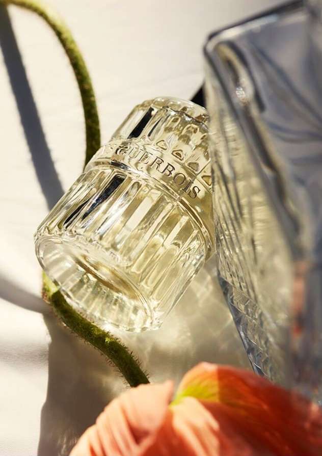Close-up of two crystal Guerbois perfume bottles next to a green stem and peach flower petal on a white surface, product shot.
