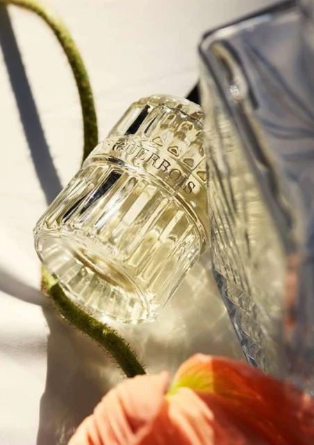 Close-up of two crystal Guerbois perfume bottles next to a green stem and peach flower petal on a white surface, product shot.