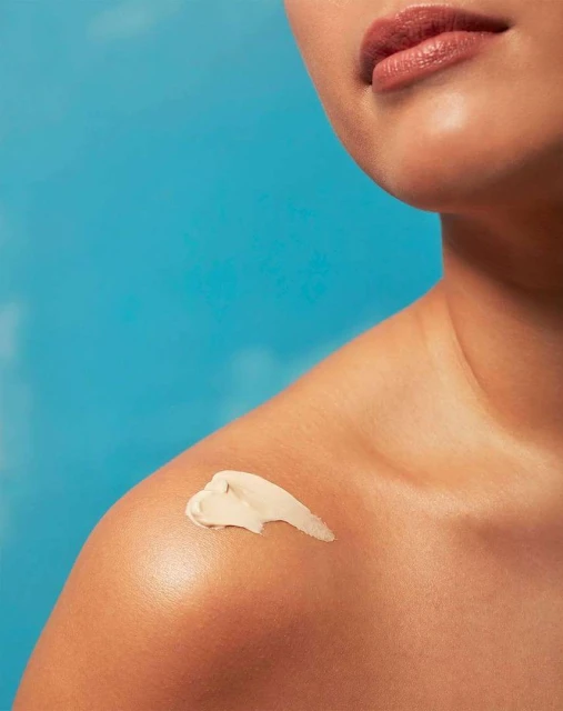 Close-up of fair-skinned woman with cream product on her shoulder against a bright blue background.