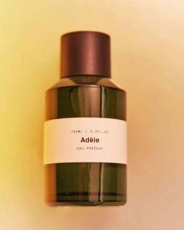 Close-up studio shot of an Adèle Eau Fraîche perfume bottle featuring a cylindrical form, dark brown cap, and a minimalist label.