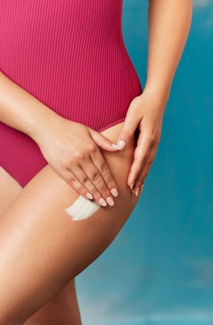 Woman in swimsuit applies lotion to her thigh in a studio setting with a blue background, focusing on skincare and body care.
