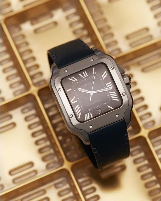 Elevated close-up studio shot of a square gray Cartier wristwatch with roman numerals and a blue strap, positioned on a golden tray.