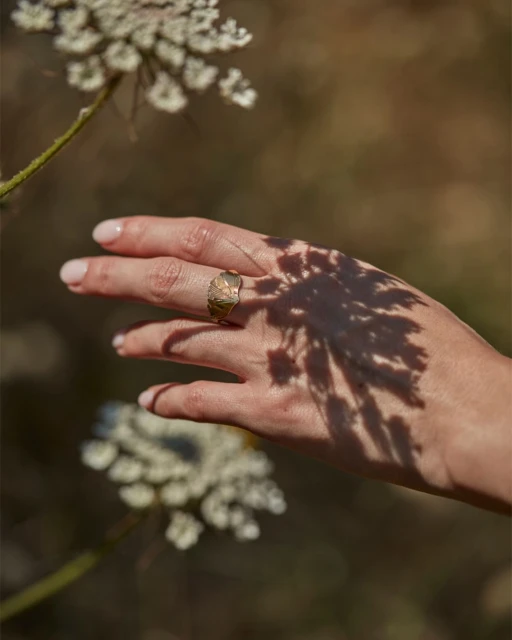 Close-up of a woman's hand with a gold ring, dappled with shadows from flowers, creating an artistic and natural jewelry display.