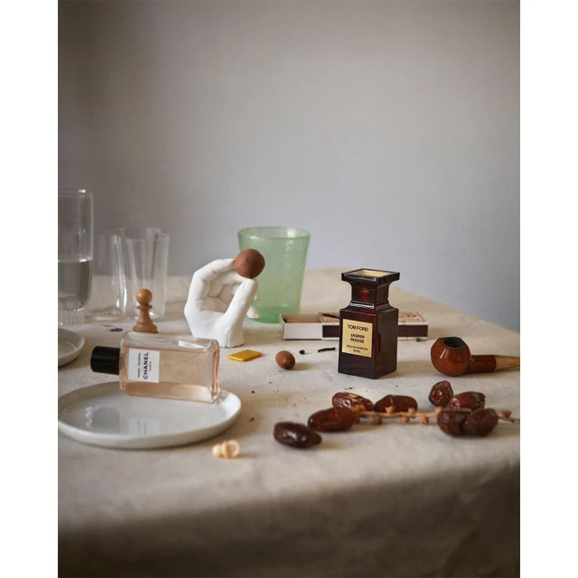 Still life tabletop arrangement of perfume bottles, glass objects, dates, and a hand sculpture on a beige tablecloth.