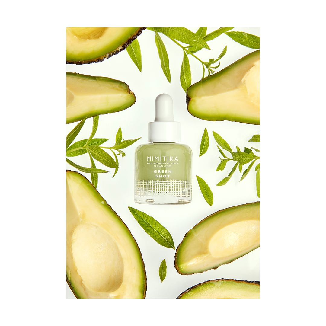 Product shot of Mimikita Green Shot serum bottle surrounded by avocado slices and green leaves, set against a white background.