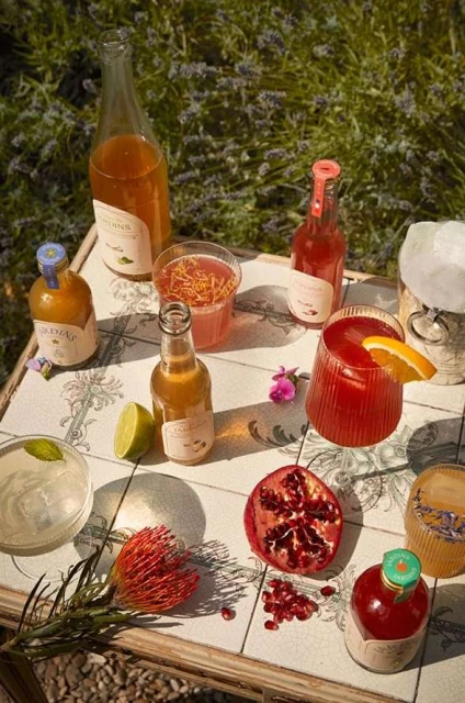 A high-angle shot displays a table laden with drinks in glass bottles and glasses, adorned with fruit, flowers, and a backdrop of lavender.