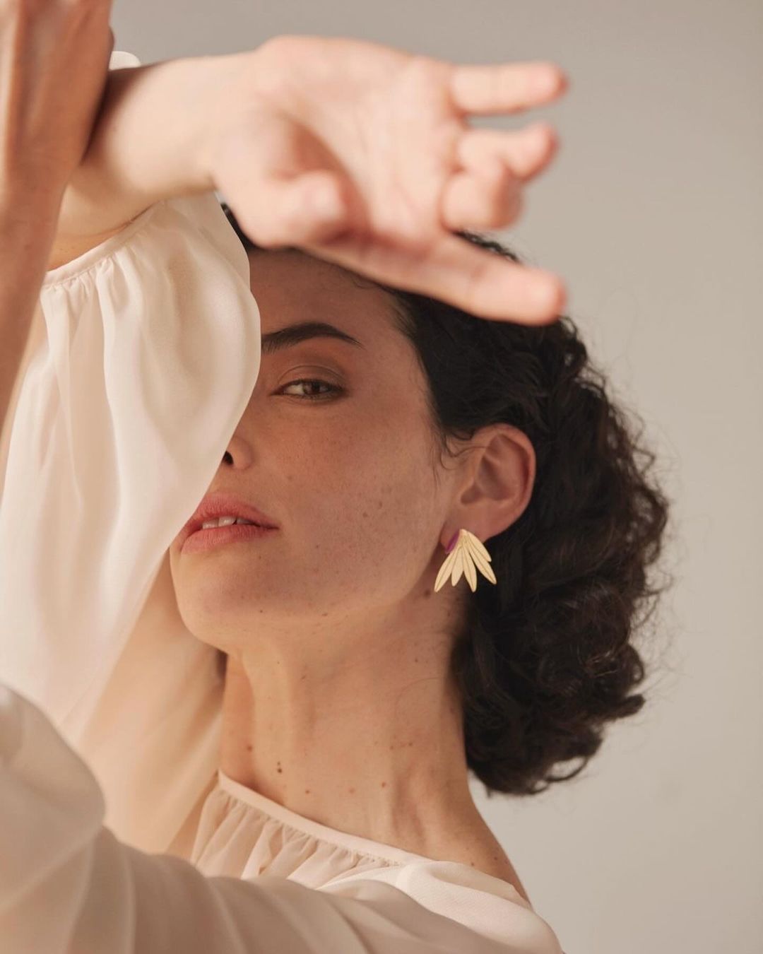 Portrait of a woman with short brown hair wearing gold earrings and a white top, with one arm raised across her face.