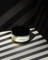 Round glass cosmetic jar with black lid and white label positioned on a surface with diagonal light and shadow stripes