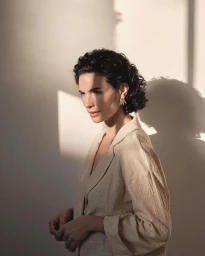 Portrait of a woman with curly dark hair wearing a beige textured jacket and leaf-shaped earrings against a plain wall with strong shadows.