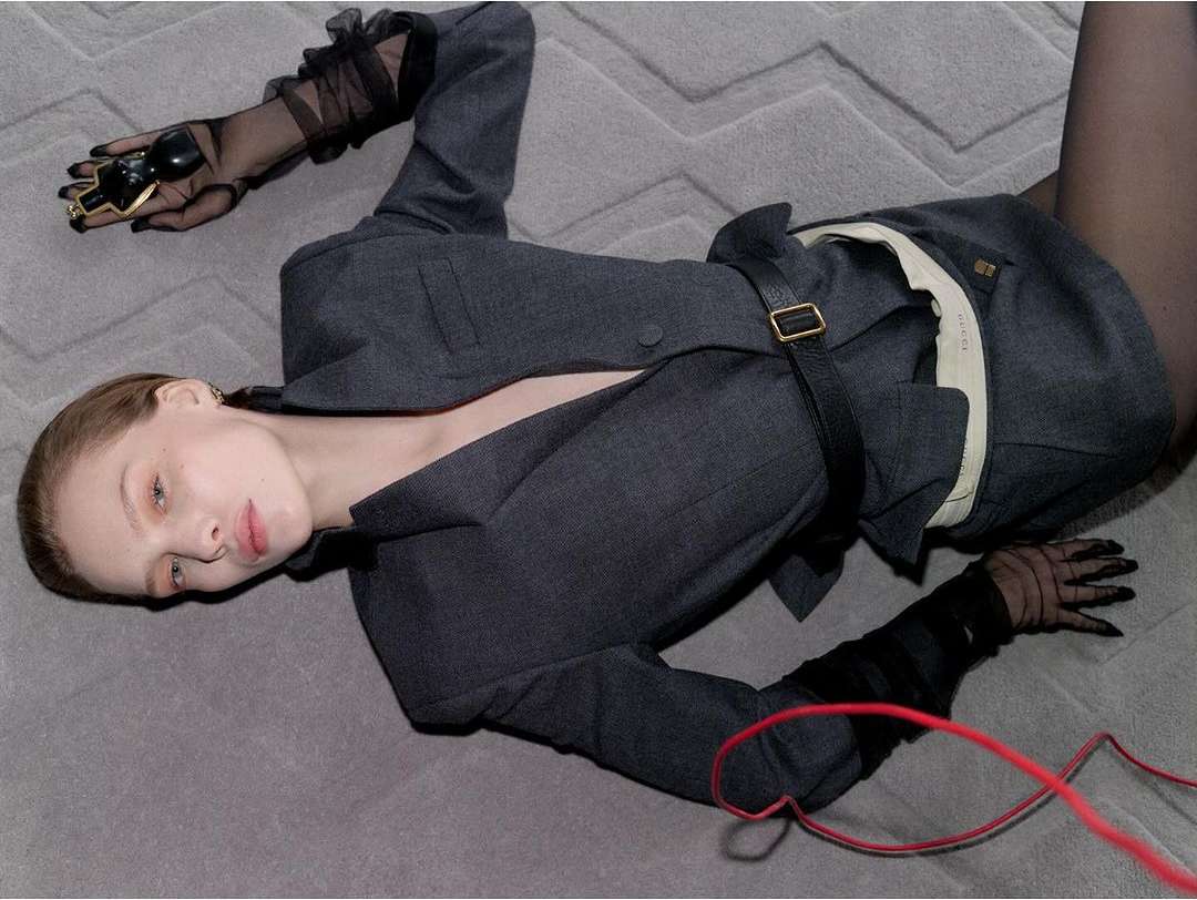 Woman in a grey suit jacket lying on a carpet, accessorized with black sheer gloves and a chunky gold ring, posing in a fashion style image.