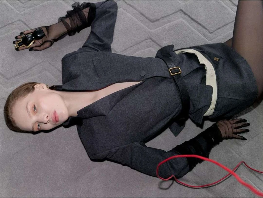 Woman in a grey suit jacket lying on a carpet, accessorized with black sheer gloves and a chunky gold ring, posing in a fashion style image.