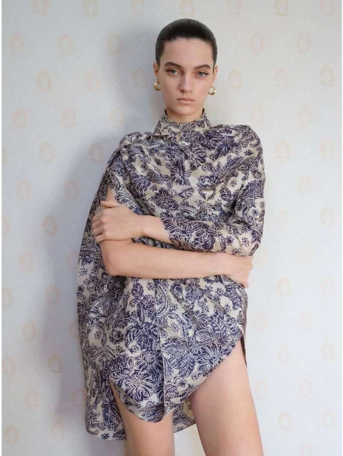 Elegant young adult woman poses indoors wearing a patterned shirt dress against a simple patterned backdrop in a fashion forward shot.