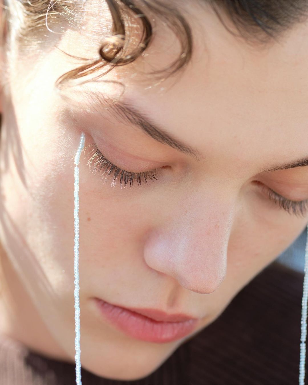 Close-up portrait of a fair-skinned young woman with light brown hair, closed eyes, and beaded strands hanging near her face.