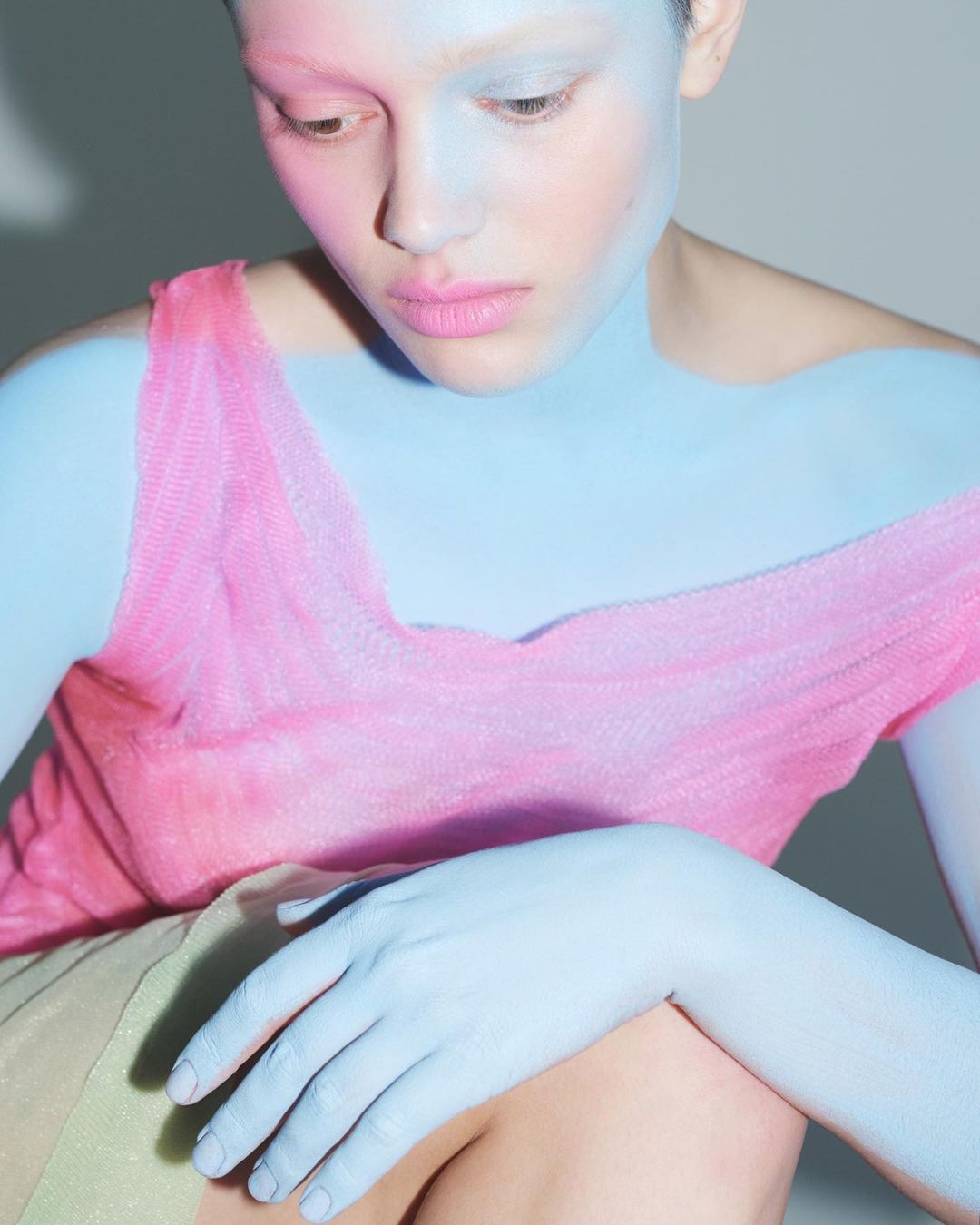 Portrait of woman with blue and pink body paint, pale skin, pink top, hand on leg, neutral expression, and light background in studio.