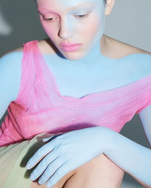 Portrait of woman with blue and pink body paint, pale skin, pink top, hand on leg, neutral expression, and light background in studio.