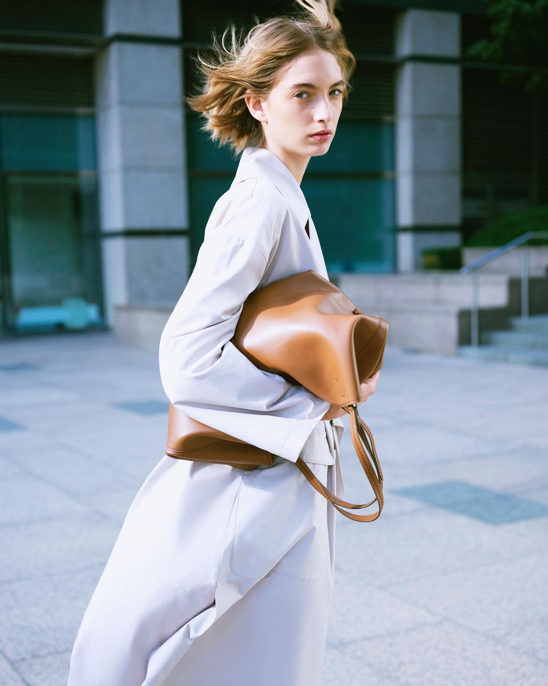 A woman in a trench coat carries a large leather bag in an urban setting, with a modern building in the background and natural light.