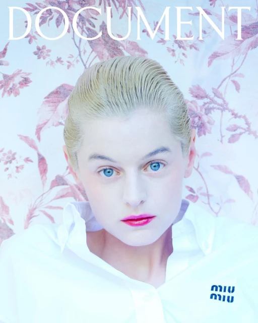 Close-up fashion shot of a fair-skinned person with platinum blonde hair and blue eyes wearing a white shirt in front of a floral background.