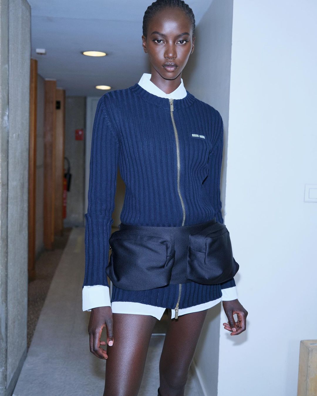 Dark-skinned woman poses indoors wearing a navy ribbed zip-up sweater with a white collared shirt and black nylon belt bag in a fashion shot.