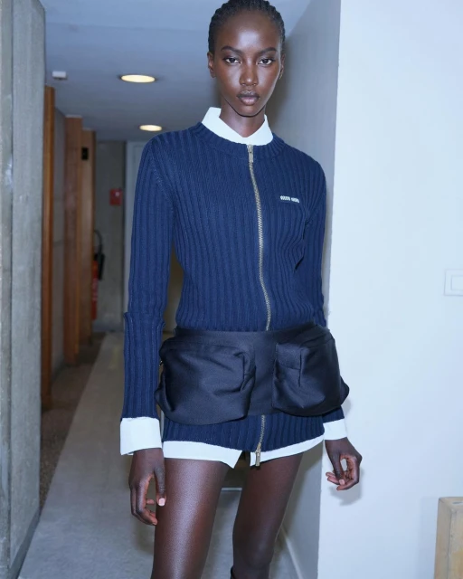 Dark-skinned woman poses indoors wearing a navy ribbed zip-up sweater with a white collared shirt and black nylon belt bag in a fashion shot.