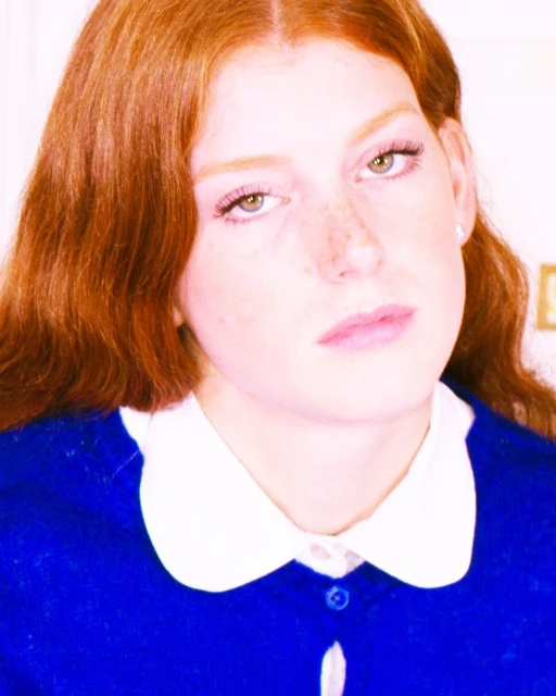Close-up portrait of a young adult female with auburn hair and light skin, wearing a blue shirt with a white collar.