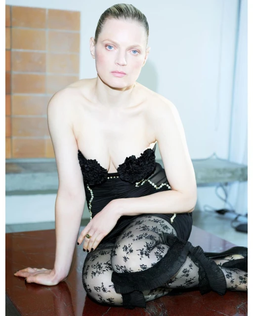A fair-skinned woman poses indoors, wearing a black strapless dress and floral lace tights, seated at a wooden table with a light background.