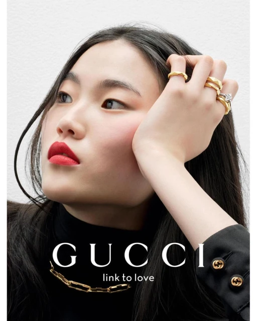 Close-up studio shot of a young woman with dark hair wearing a black turtleneck and gold necklace, adorned with gold rings on her fingers.