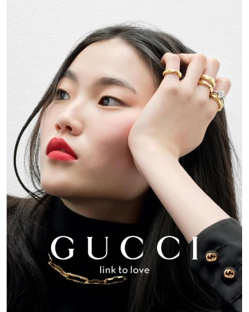 Close-up studio shot of a young woman with dark hair wearing a black turtleneck and gold necklace, adorned with gold rings on her fingers.
