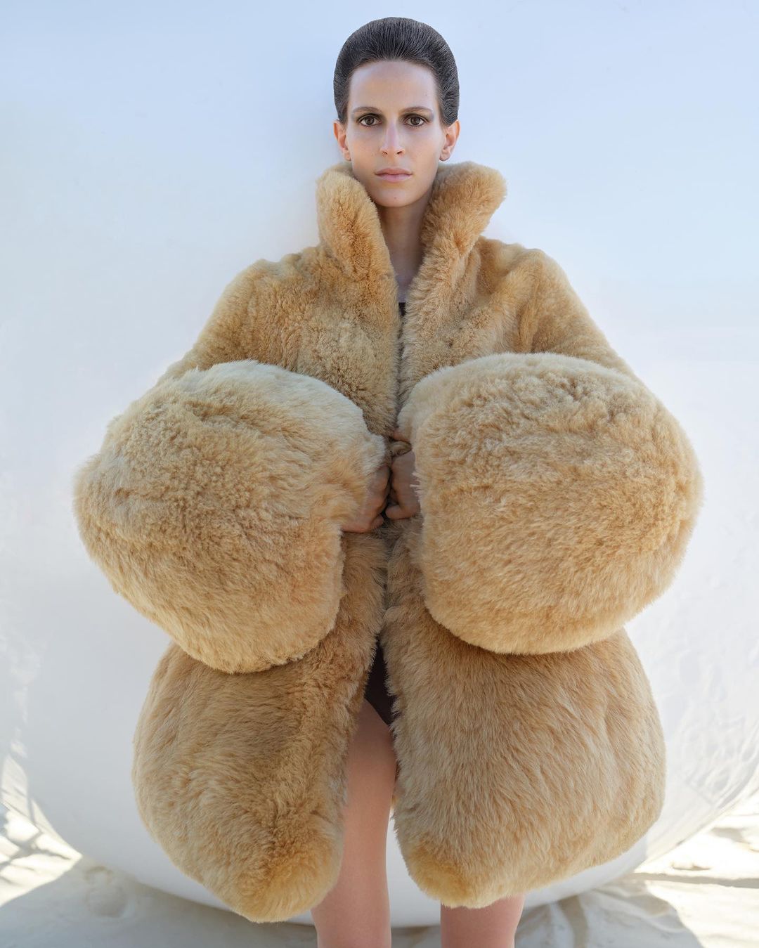 Fashion shot of a model posing in a beige oversized fur coat with large sleeves in front of a white wall, focusing on texture and design.