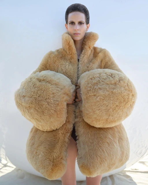 Fashion shot of a model posing in a beige oversized fur coat with large sleeves in front of a white wall, focusing on texture and design.