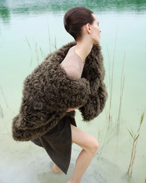 Woman wearing a fur coat and skirt stands in a shallow, light-green body of water surrounded by tall grasses in an outdoor setting.