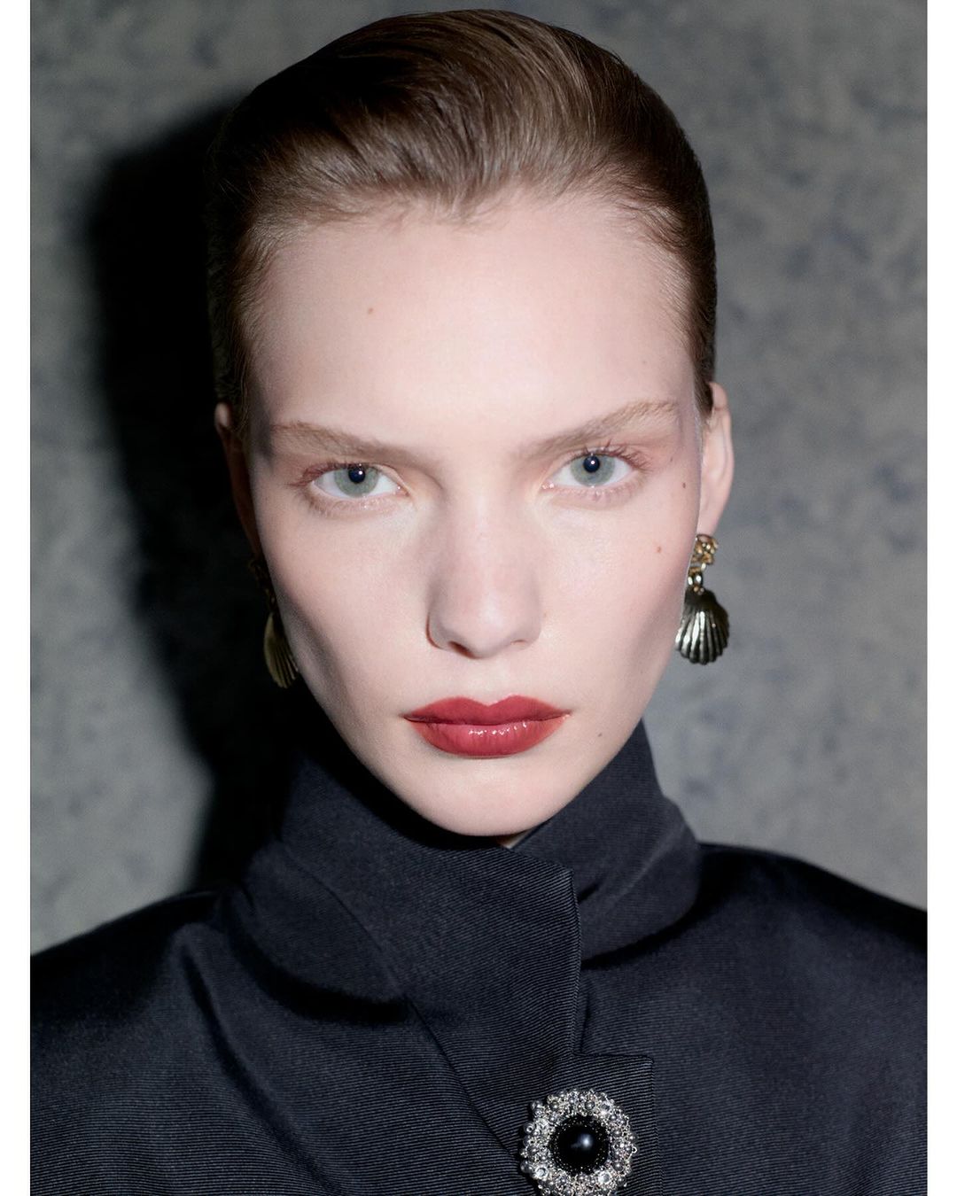 Close-up studio portrait of a fair-skinned woman with red lipstick, wearing dark coat with ornate brooch and shell earrings against grey backdrop.