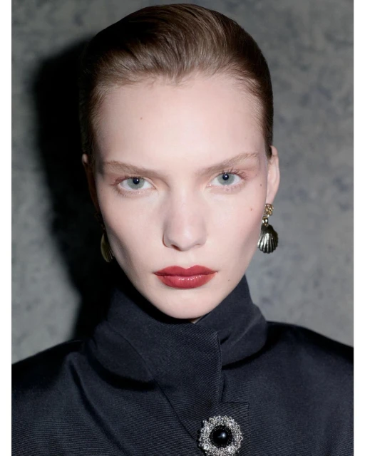 Close-up studio portrait of a fair-skinned woman with red lipstick, wearing dark coat with ornate brooch and shell earrings against grey backdrop.
