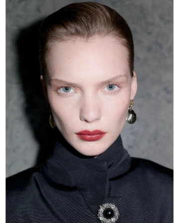 Close-up studio portrait of a fair-skinned woman with red lipstick, wearing dark coat with ornate brooch and shell earrings against grey backdrop.
