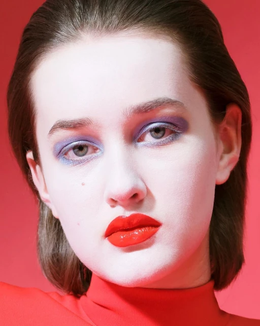Close-up portrait of a young adult female with short brown hair, red turtleneck, and bold purple eyeshadow and red lipstick against pink backdrop.