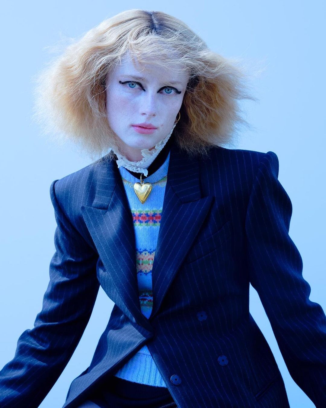 A young female model with blonde hair wears a pinstripe blazer over a blue fair isle sweater and gold heart necklace against a light blue backdrop.
