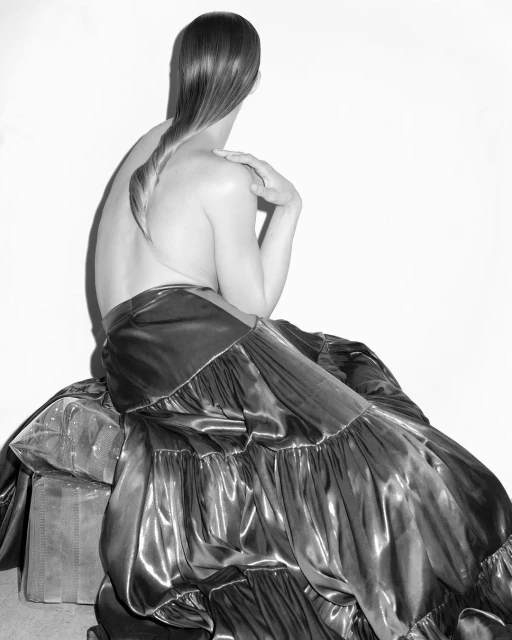 Black and white portrait of an adult with a long braid sitting on luggage, looking away from the camera with a light background.