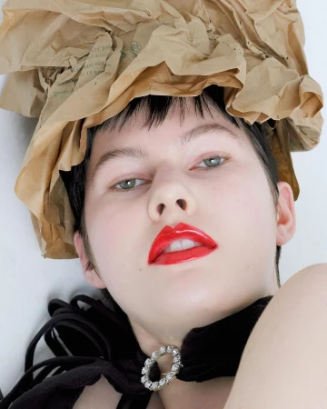 Close-up studio portrait of a woman with pale skin, red lips, and paper bag headwear against a white background.