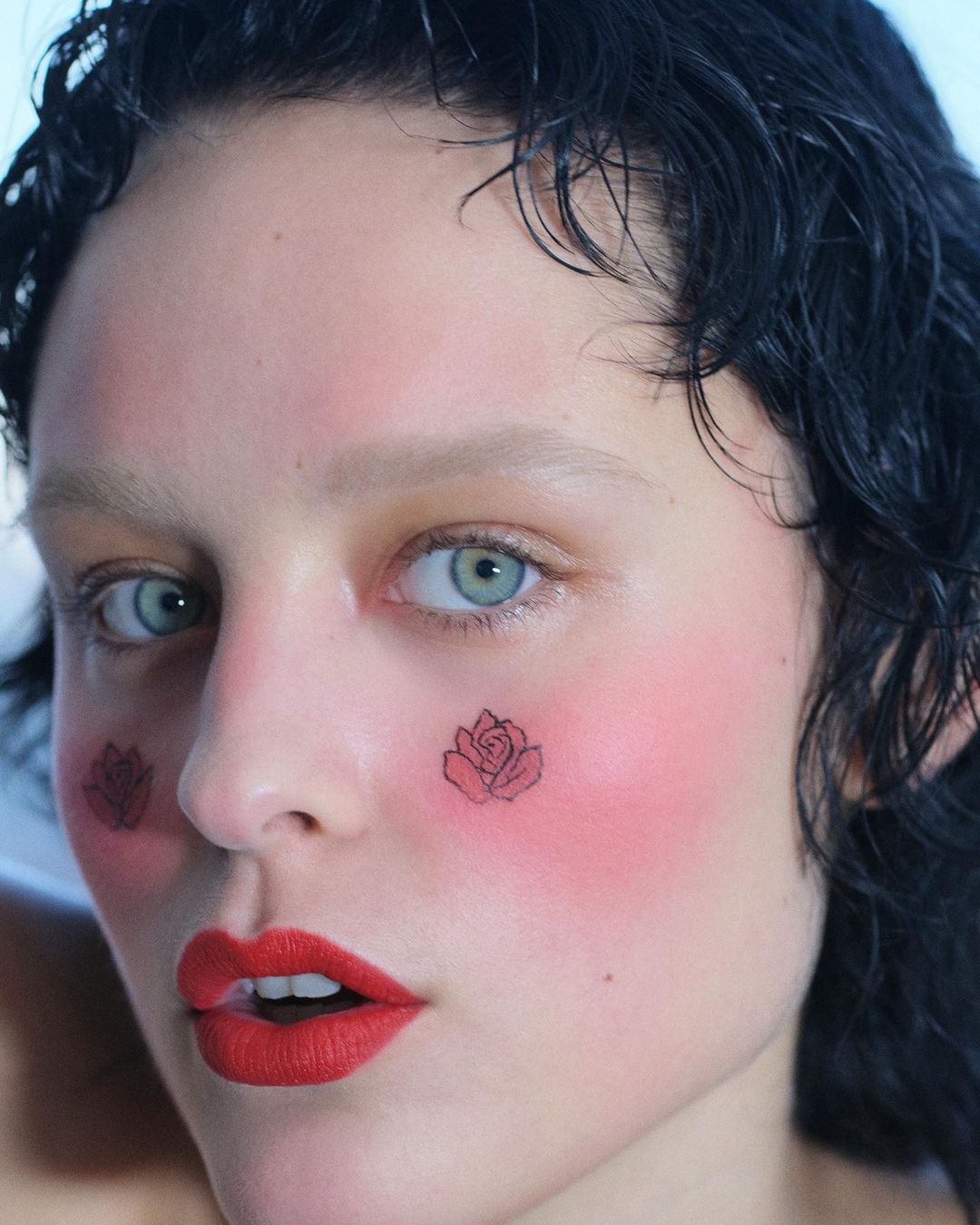Close-up portrait of a fair-skinned person with red lipstick, rose tattoos on their face, and wet curly black hair.