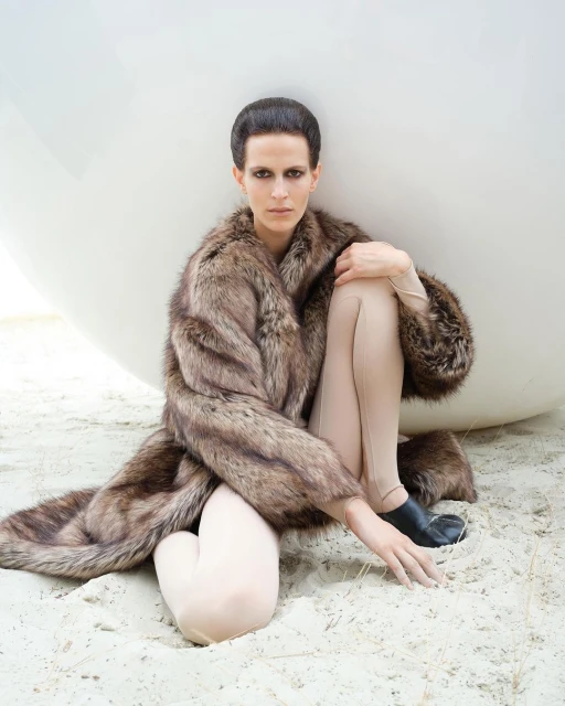 A woman poses on a sandy surface wearing a long, patterned fur coat with a neutral top and leggings, positioned near a large white sphere.