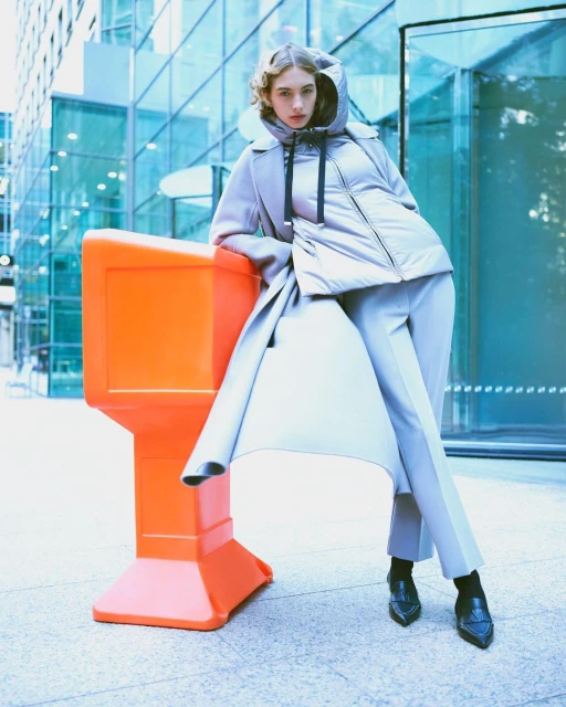 Fashionable woman in winter outerwear poses outdoors in city against a modern building, leaning against an orange traffic barrier.